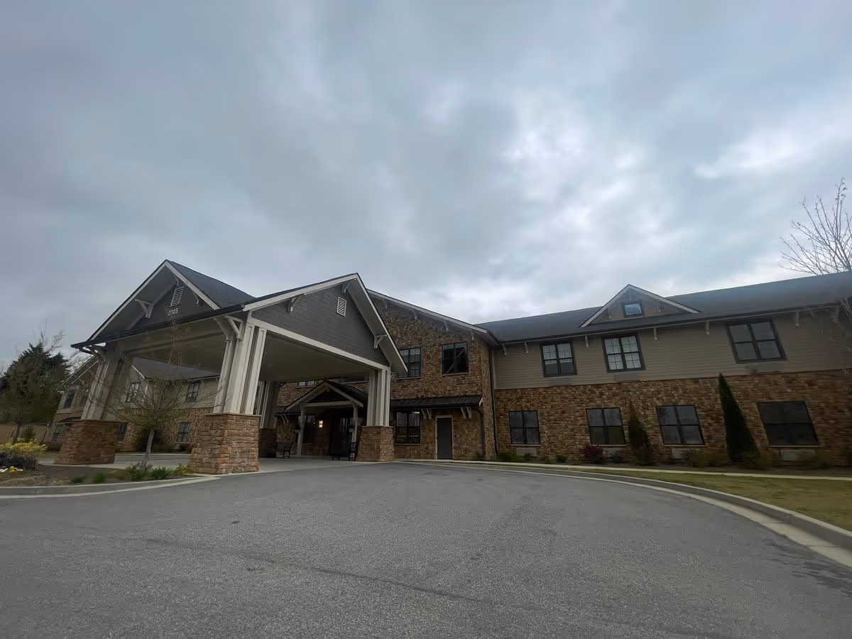 Front exterior of a two-story senior living building with a covered porte-cochère and stone facade under an overcast sky.