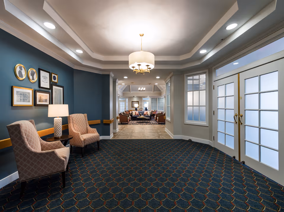 A well-lit interior hallway in a senior living facility with patterned carpet, two upholstered chairs and a side table with a lamp on the left, framed artwork on the wall, and double glass doors on the right. The hallway leads to a spacious common area with multiple seating arrangements and large windows.