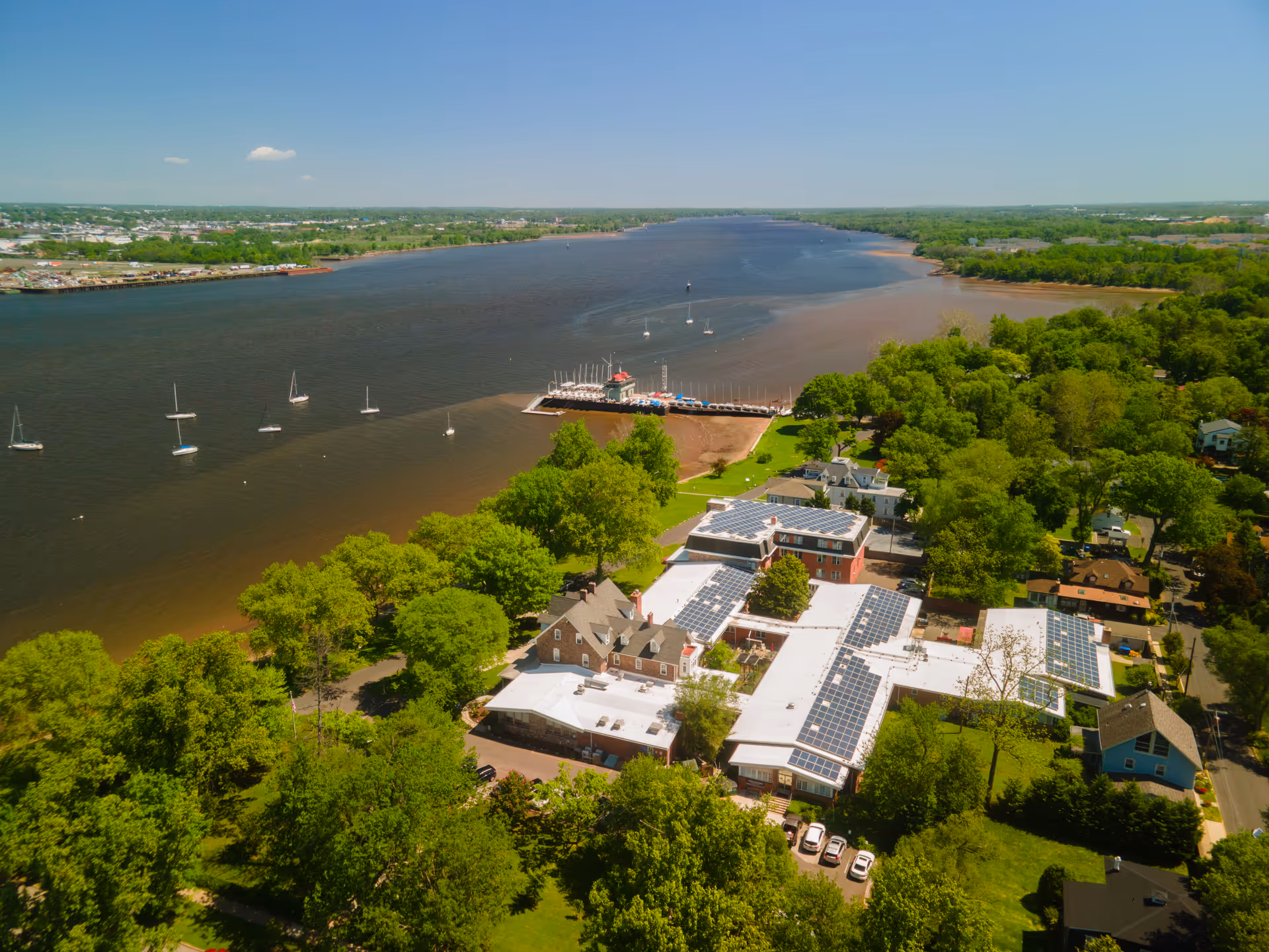 Aerial view of Riverview Estates facility near a large river with several sailboats anchored. The facility buildings have solar panels on the roofs and are surrounded by lush green trees and residential houses. The river extends into the horizon under a clear blue sky.
