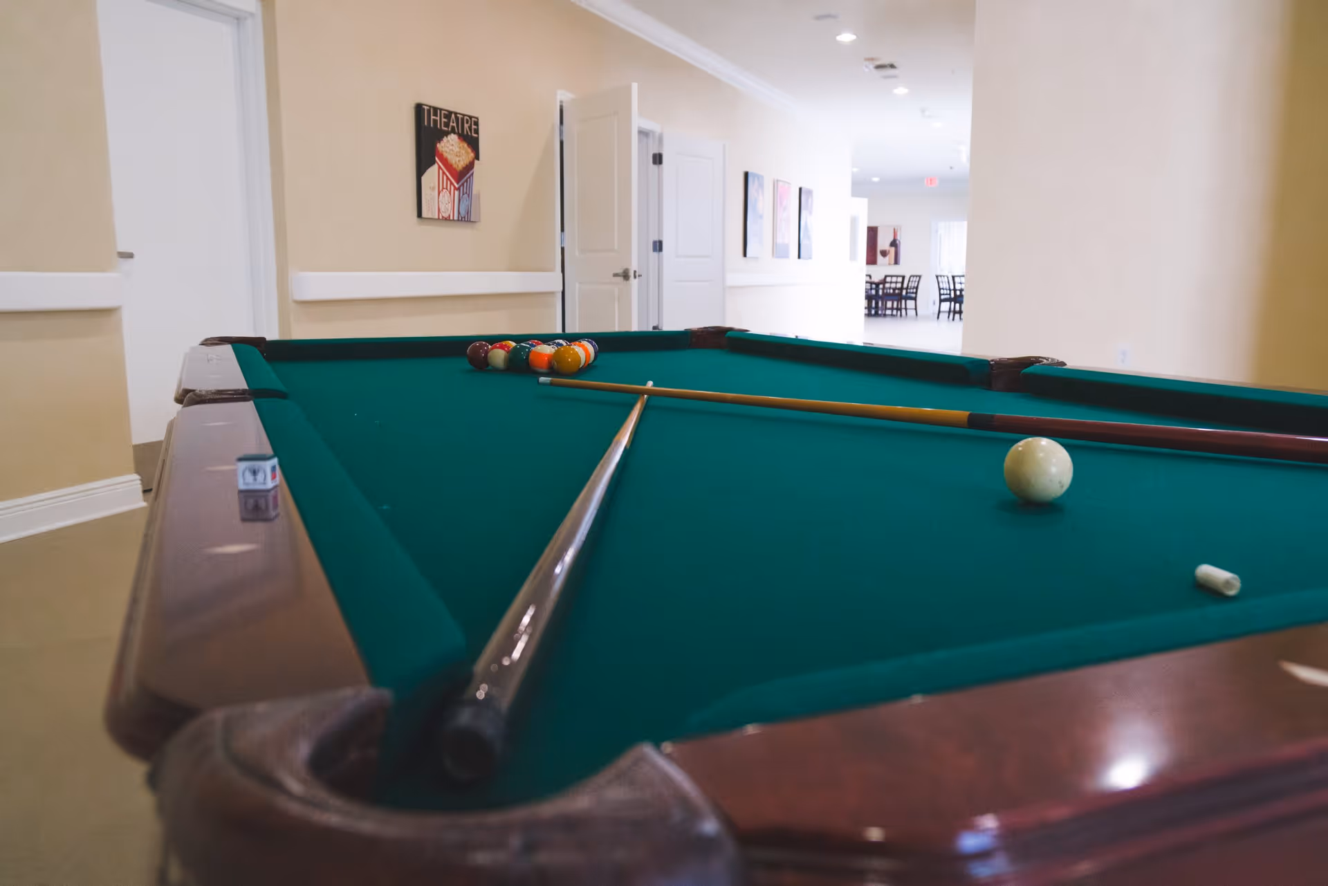 A green felt pool table with billiard balls arranged in a triangle and two pool cues resting on the table in a well-lit hallway with beige walls and white doors. A sign on the wall reads 'THEATRE' with a popcorn image, and there are chairs and tables visible in the background.