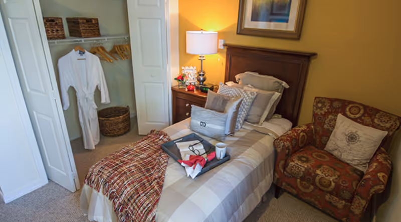A cozy bedroom with a single bed covered in beige and white checkered bedding, multiple pillows, and a red patterned throw blanket. A tray with a book, glasses, a napkin, and a cup is placed on the bed. Next to the bed is a wooden nightstand with a lamp, flowers, and decorative items. An armchair with a patterned fabric and a pillow is positioned beside the bed. In the background, there is an open closet with a white robe hanging and some baskets on the shelf.