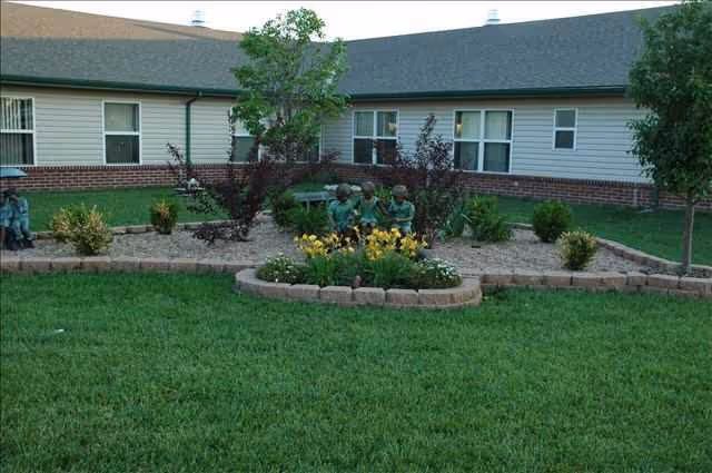 A landscaped garden area with green grass, flower beds bordered by stone edging, small trees, and shrubs in front of a single-story building with beige siding and brick trim. There are three small statues of children sitting on a bench in the garden.