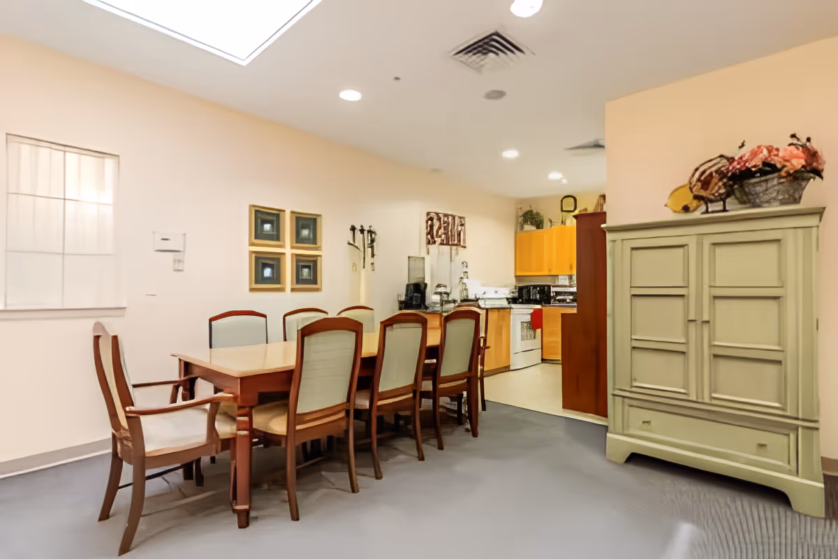 Dining room with a long wooden table surrounded by chairs, an open kitchen in the background, and a green storage cabinet.