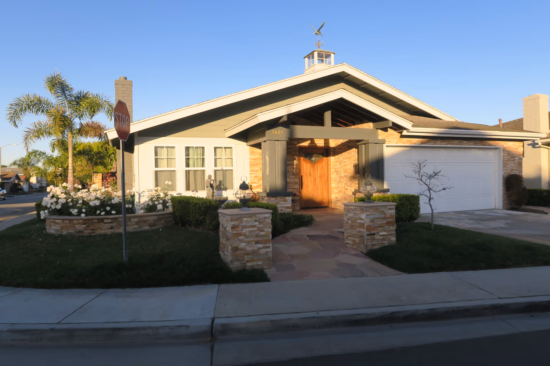 Exterior view of a single-story residential building with a front porch, a wooden door, a garage, and a well-maintained garden with white flowers and palm trees. A stop sign is visible on the sidewalk near the corner of the property.