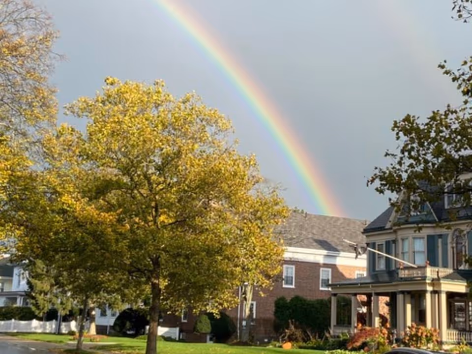 A rainbow arches over trees and a Victorian-style house with a front porch.