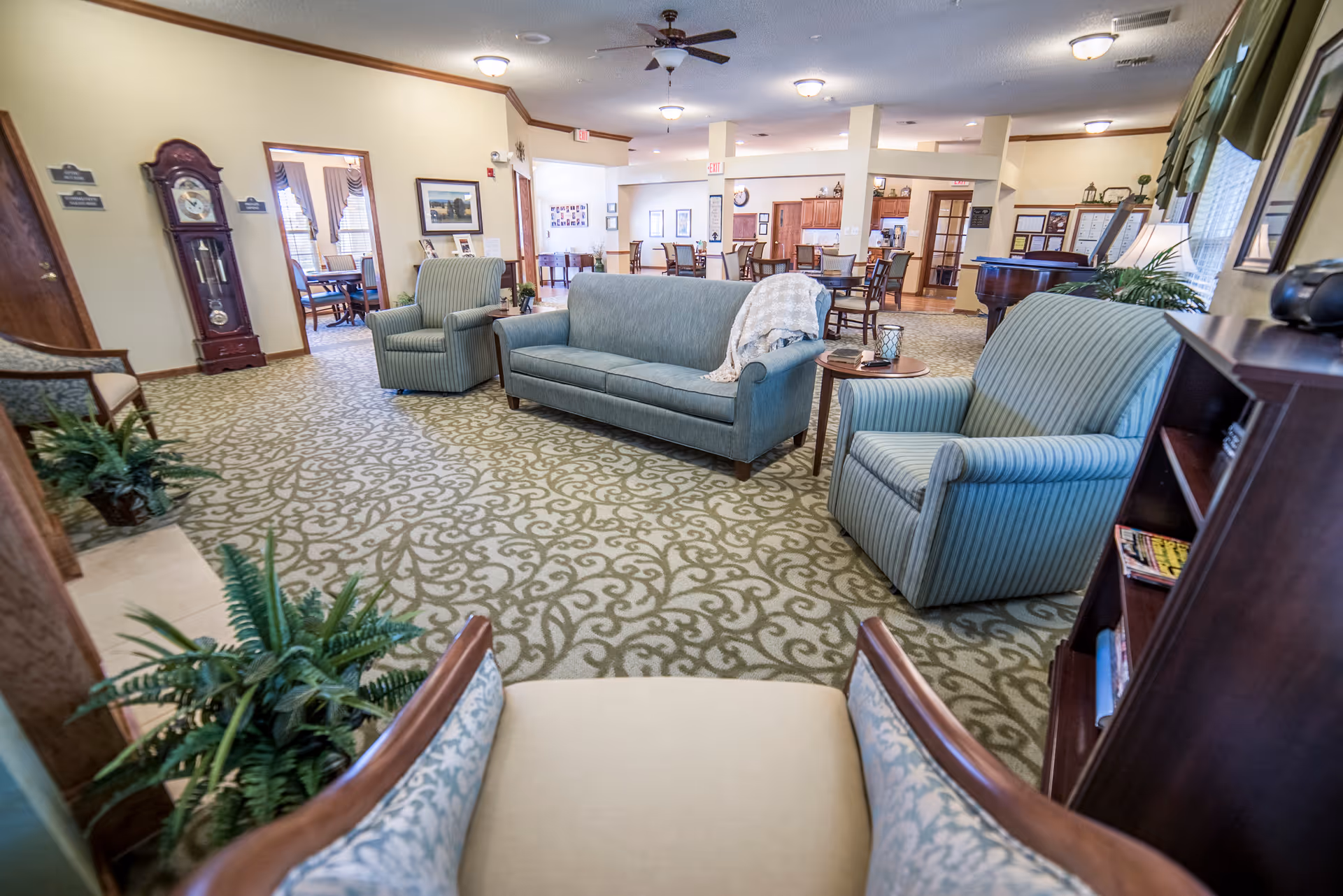 A bright senior living facility common lounge with sofas, armchairs, patterned carpet and a dining area visible in the background.