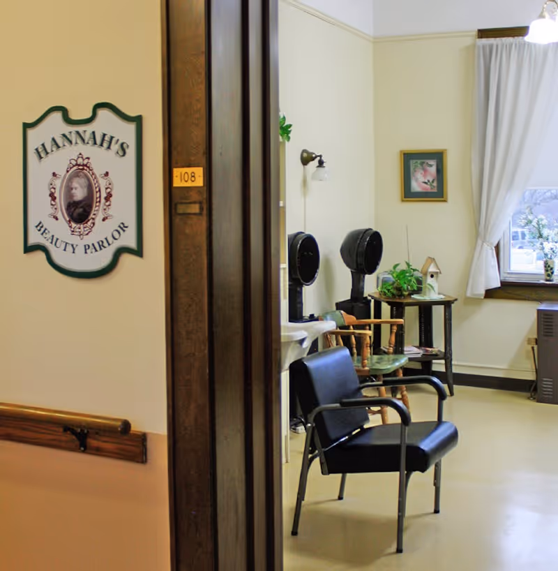 Interior view of a beauty parlor room named Hannah's Beauty Parlor. The room contains black salon chairs, hair drying stations, a small table with plants and a birdhouse, a window with white curtains, and a framed picture on the wall.