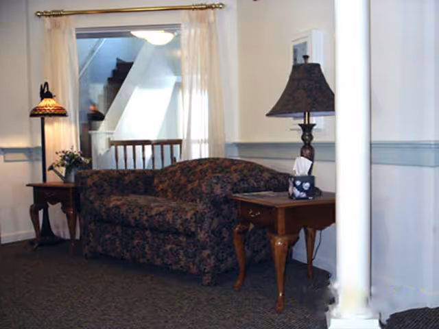 Interior view of a living room area featuring a floral patterned sofa between two wooden side tables. Each table has a lamp, one with a stained glass shade and the other with a dark fabric shade. A window with sheer curtains and a staircase are visible in the background.
