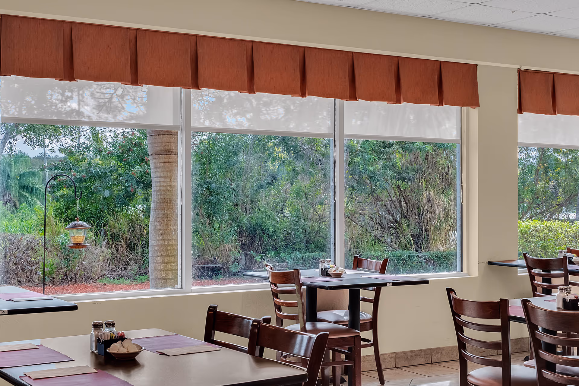 Dining area with several tables and wooden chairs set near large windows showing a green outdoor garden. Tables have placemats, salt and pepper shakers, and small condiment containers. The windows have white roller shades and red valances.