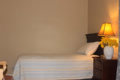 A simple bedroom with a single bed covered in a striped bedspread, a pillow, a wooden nightstand with drawers, a white vase with yellow flowers, and a lit table lamp.