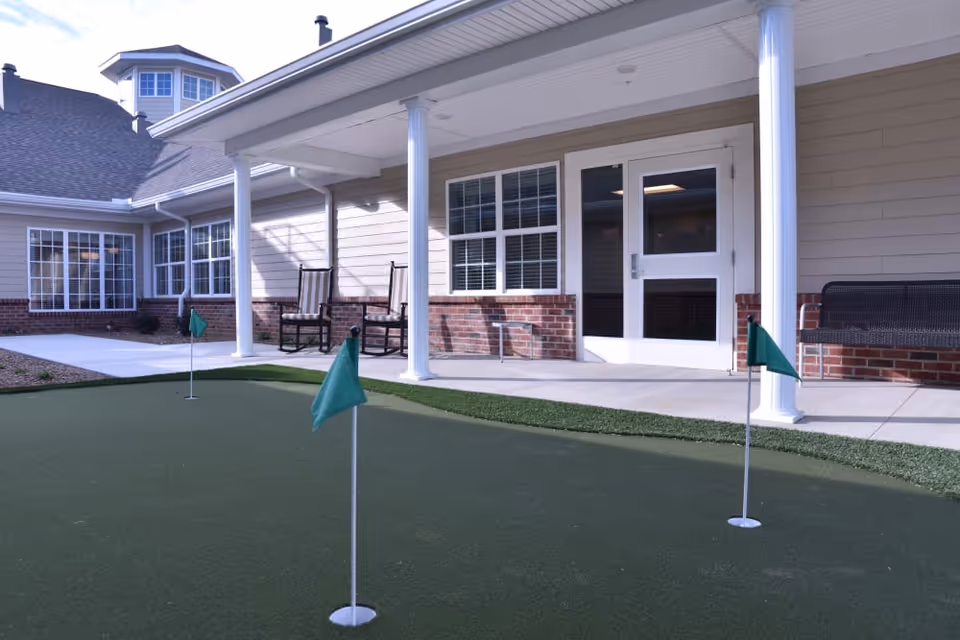 Outdoor putting green with three small green flags in front of a building with beige siding, white columns, and large windows. There are two rocking chairs and a bench on the covered patio area.