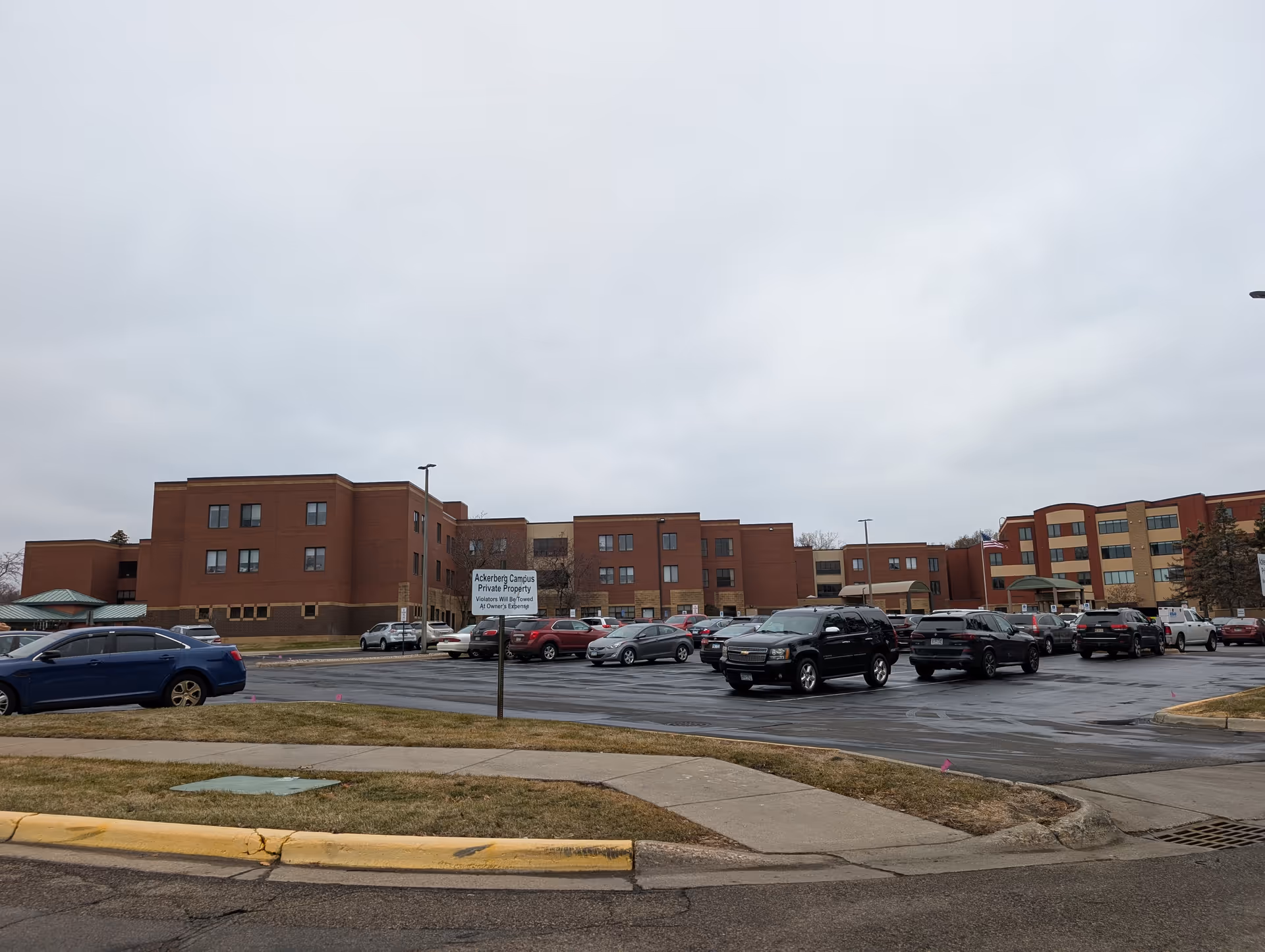 Parking lot with cars in front of a multi-story brick assisted living building under an overcast sky.