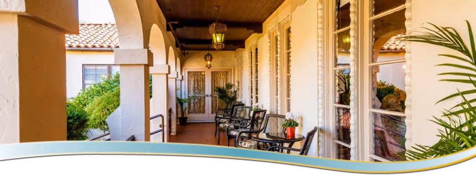 Covered arched porch with seating and small tables along a walkway beside windows.