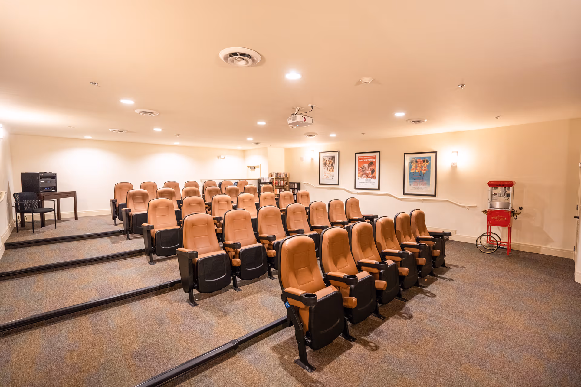 A small theater room with three rows of tan cushioned seats arranged on tiered carpeted flooring. The walls are light-colored with three framed movie posters on one side. There is a red popcorn machine on the right side near the wall and a small table with audio equipment and a chair on the left side. Ceiling lights and a projector are visible overhead.