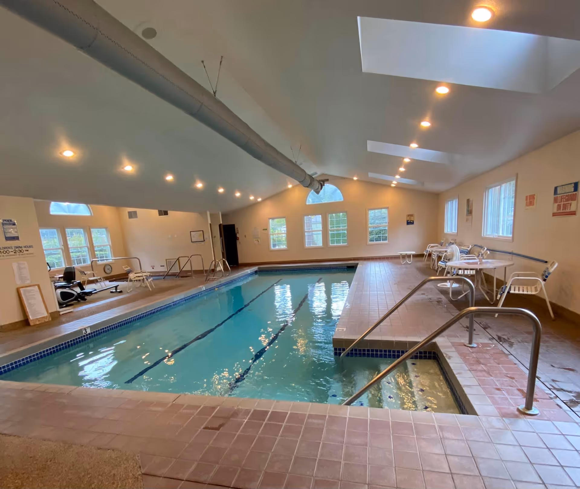 Indoor swimming pool with tiled deck, metal handrails, and several chairs and tables along the side. The room has multiple windows, skylights, and ceiling lights, with exercise equipment visible in the background.