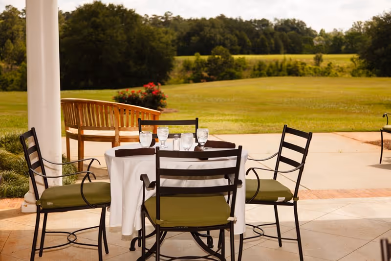 Outdoor patio area with a round table covered with a white tablecloth, set with four glasses of water and brown napkins. Four black metal chairs with green cushions surround the table. In the background, there is a wooden bench, green grass, bushes, and trees under a partly cloudy sky.