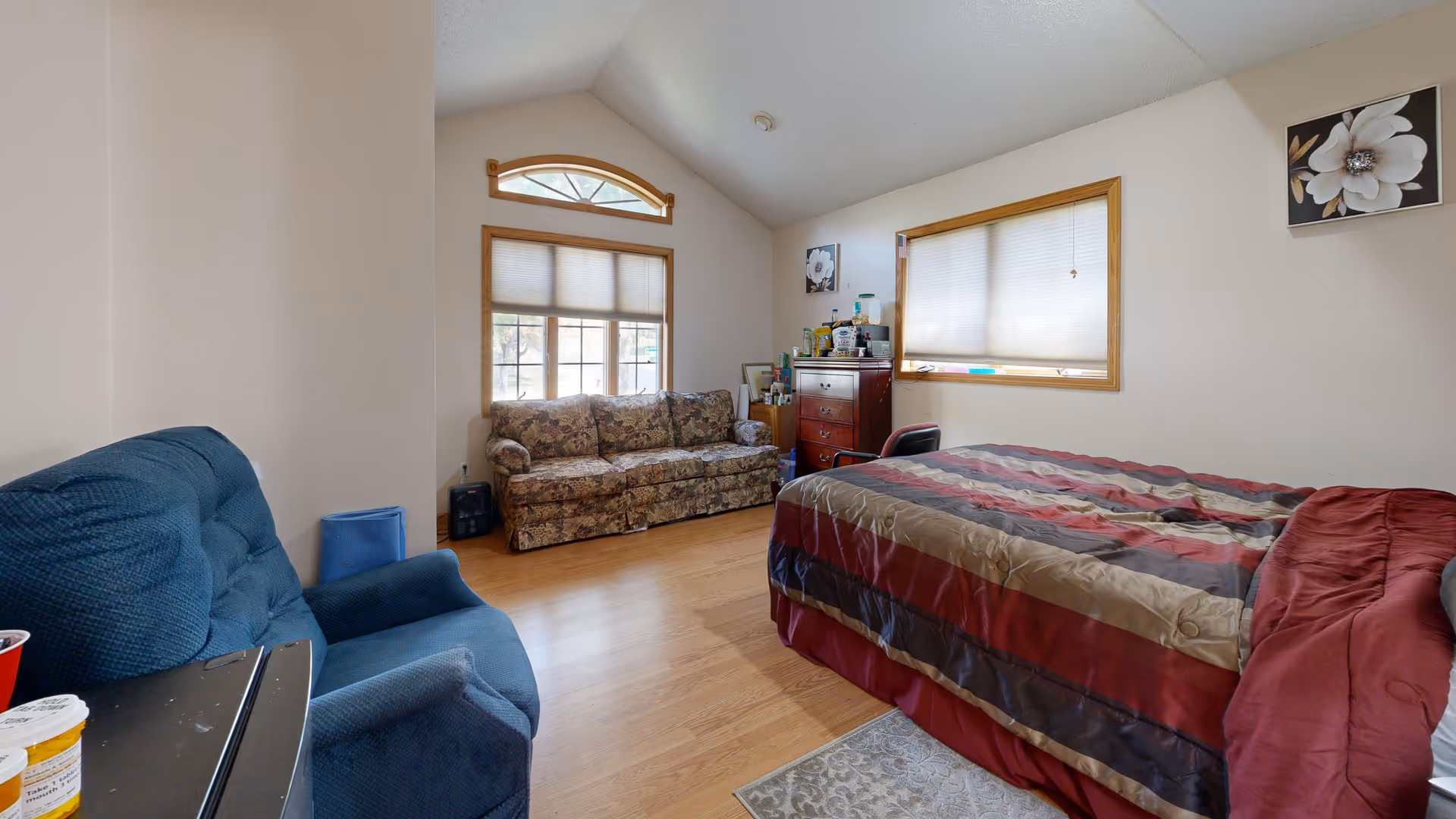 A bedroom with a large bed covered in a striped comforter, a blue armchair, a floral-patterned sofa, a wooden dresser with various items on top, and two windows with blinds partially closed. The room has light-colored walls and a wooden floor.