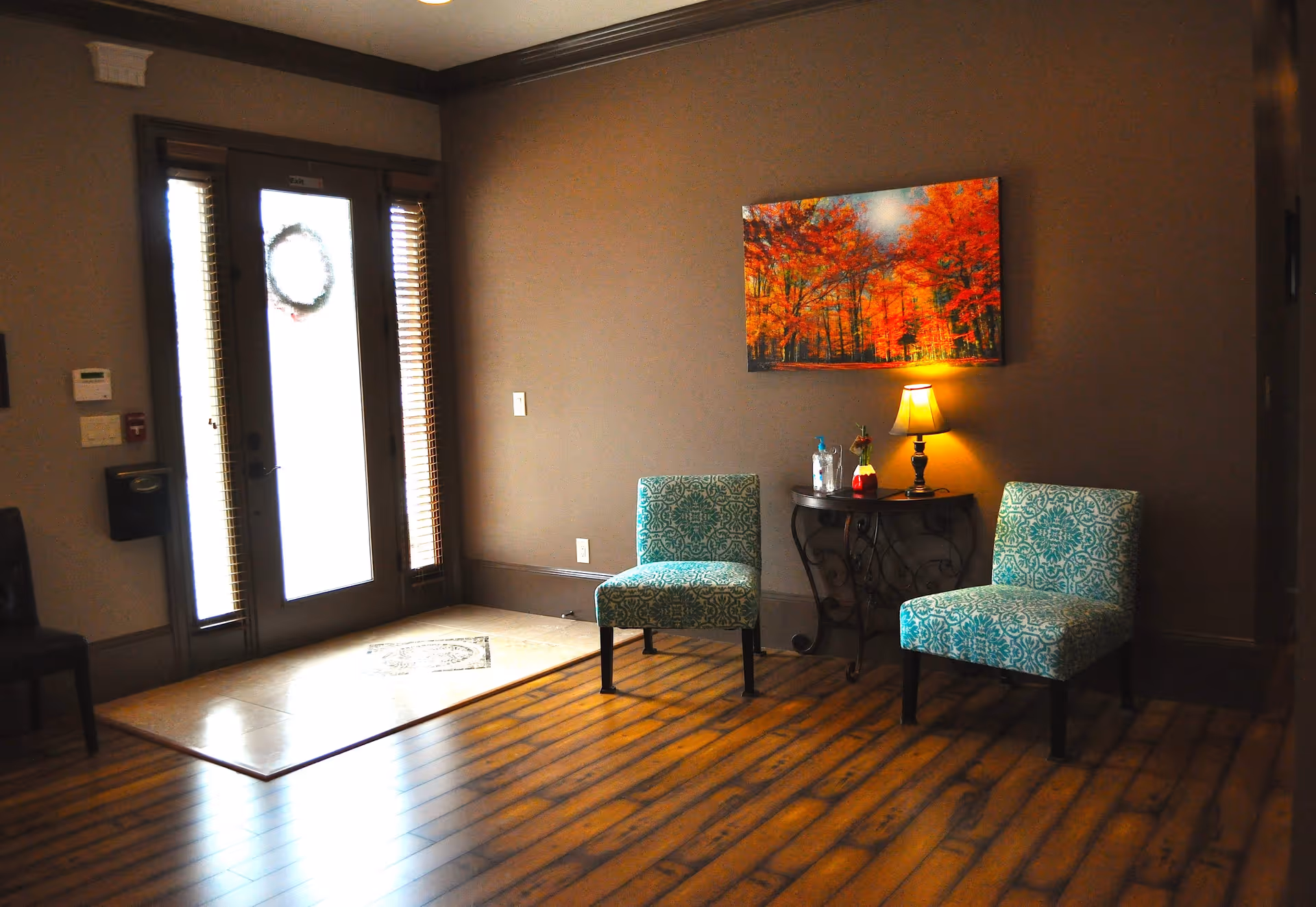 Interior view of a senior living facility entrance area with two patterned teal chairs, a small round table with a lamp and decorative items, wooden flooring, and a door with frosted glass panels and a wreath. A colorful autumn forest painting hangs on the wall.