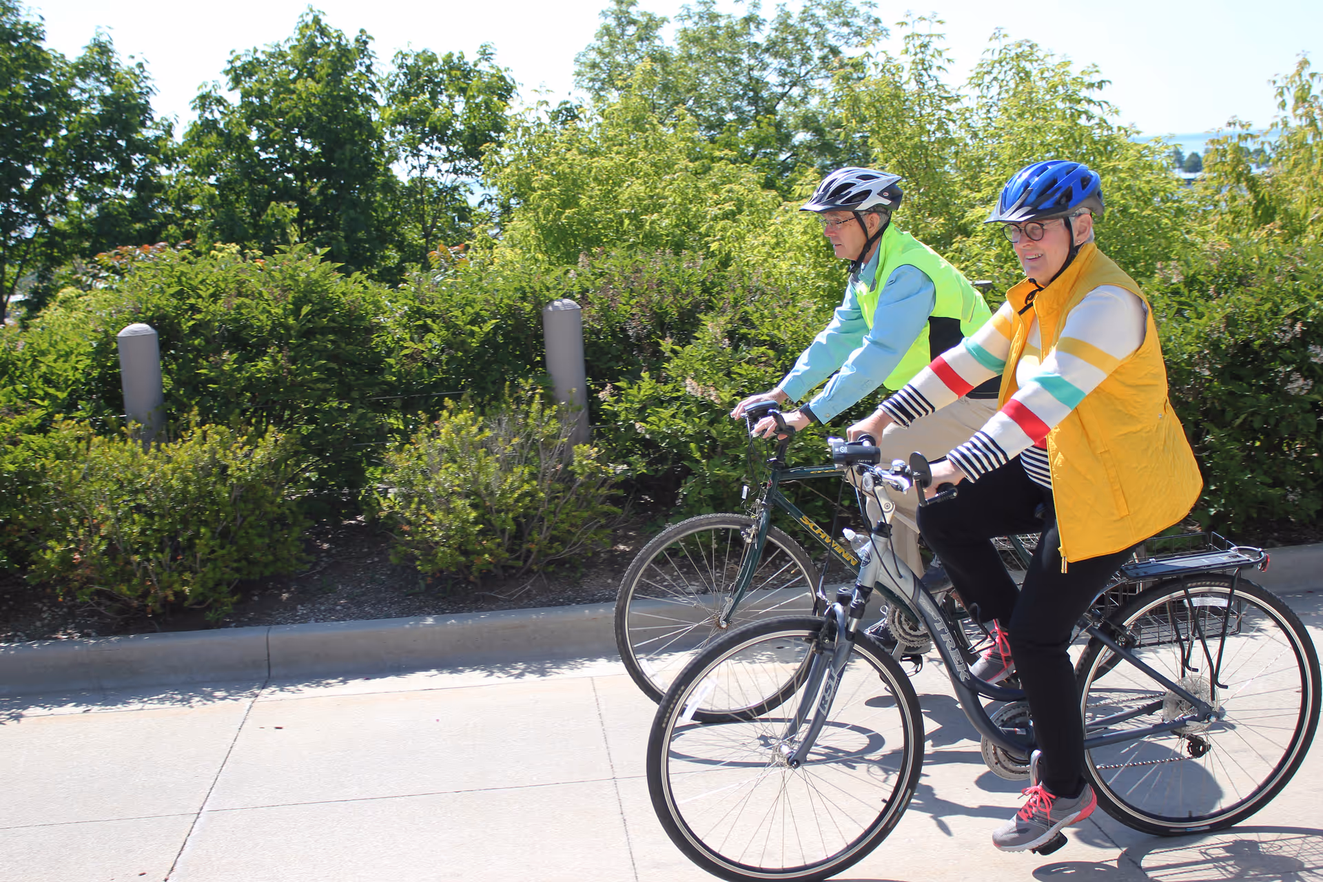 Two elderly individuals wearing helmets and casual clothing riding bicycles on a paved path with green bushes and trees in the background on a sunny day.