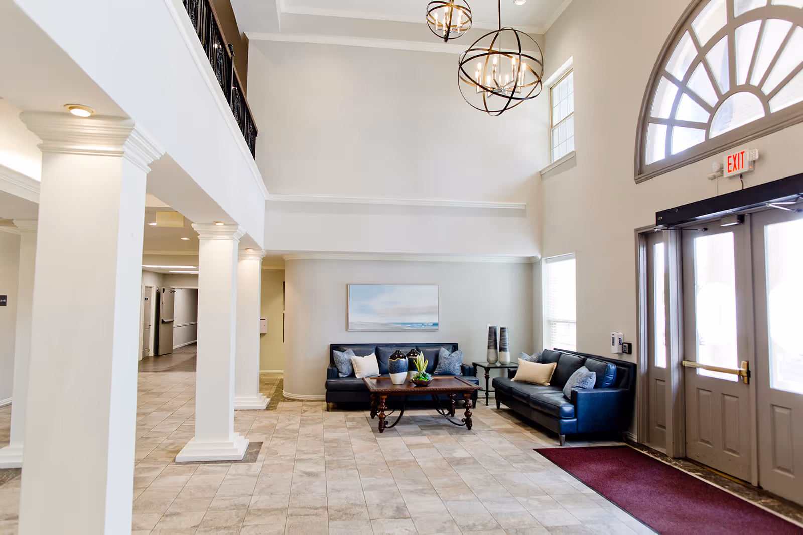 A bright and spacious lobby area with high ceilings, featuring two black leather sofas with cushions, a wooden coffee table with decorative plants, large windows, and an arched window above the entrance door. The floor is tiled, and there are white columns supporting the ceiling.