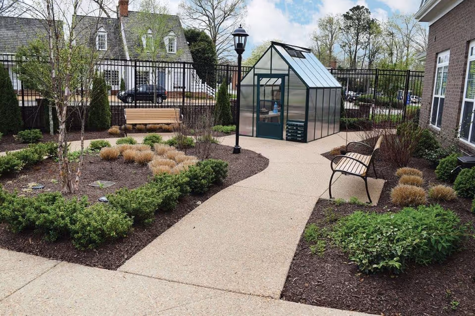 Courtyard garden with a paved walkway, benches, a small greenhouse and landscaped plantings beside a building.