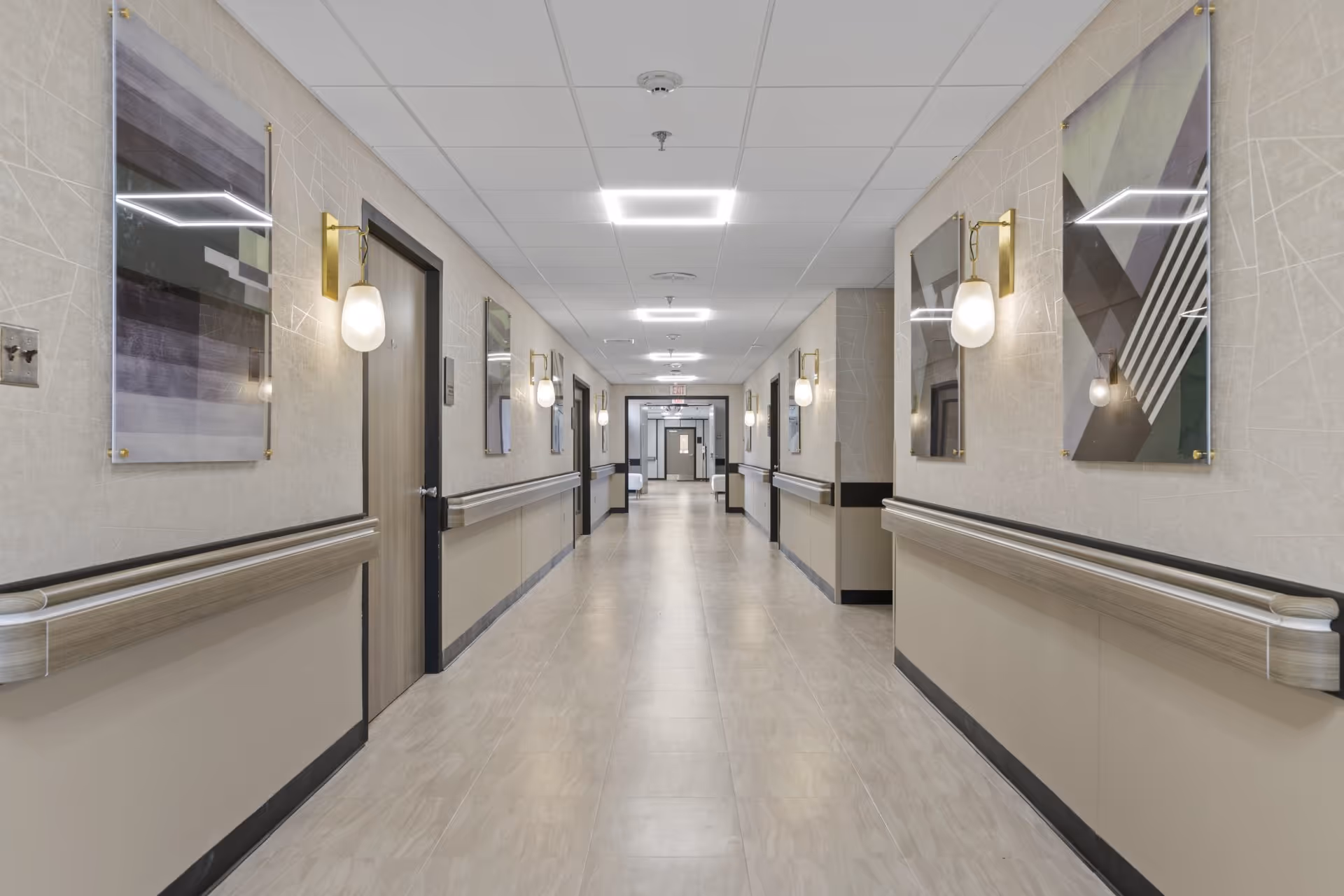A long, clean, and well-lit hallway in a senior living facility with beige walls, light-colored tiled floors, and multiple closed doors on both sides. The walls are decorated with modern abstract artwork and wall-mounted light fixtures with frosted glass shades. Handrails run along both sides of the hallway for support.