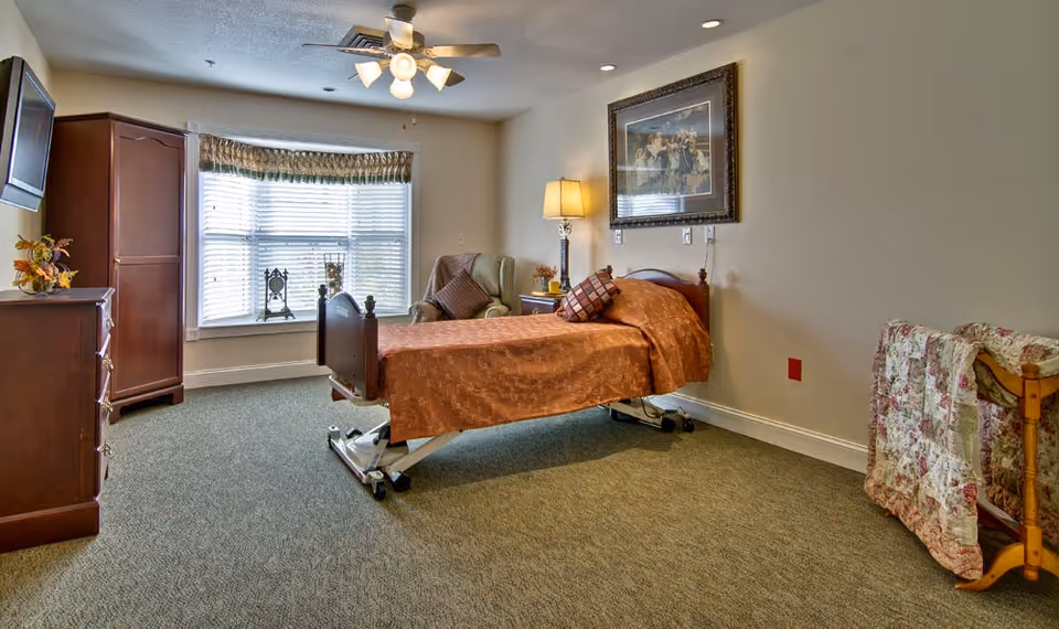 A furnished bedroom with a single adjustable bed, armchair, dresser, wall-mounted TV, and a large bay window.