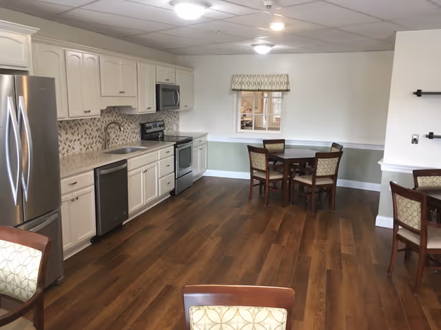 Communal kitchen and dining area with white cabinetry, stainless steel appliances, wood floors, and several dining tables and chairs.