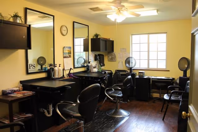 Interior view of a hair salon area with black styling chairs, mirrors on the wall, hair washing sinks, and shelves with hair care products. The room has yellow walls, a ceiling fan with lights, and a window letting in natural light.