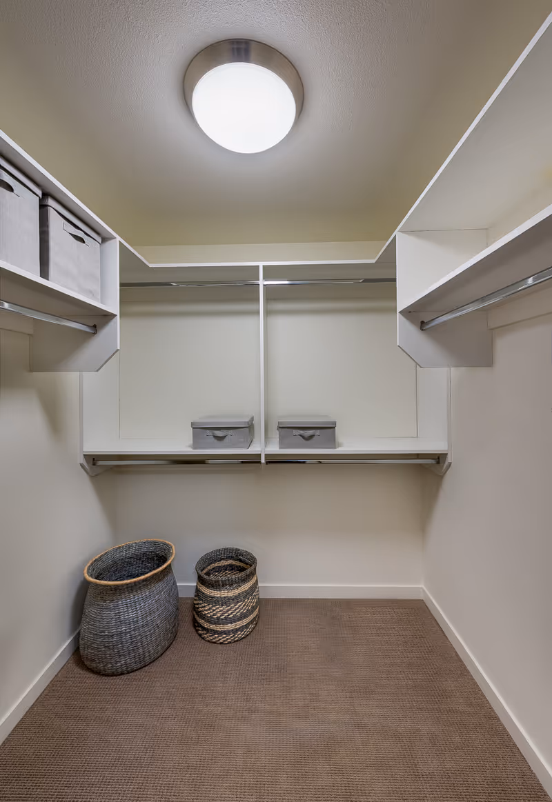 A walk-in closet with beige walls and carpeted floor. The closet has white shelving units with hanging rods on both sides and at the back. There are two gray storage boxes on the back shelf and two woven baskets on the floor. A round ceiling light fixture illuminates the space.