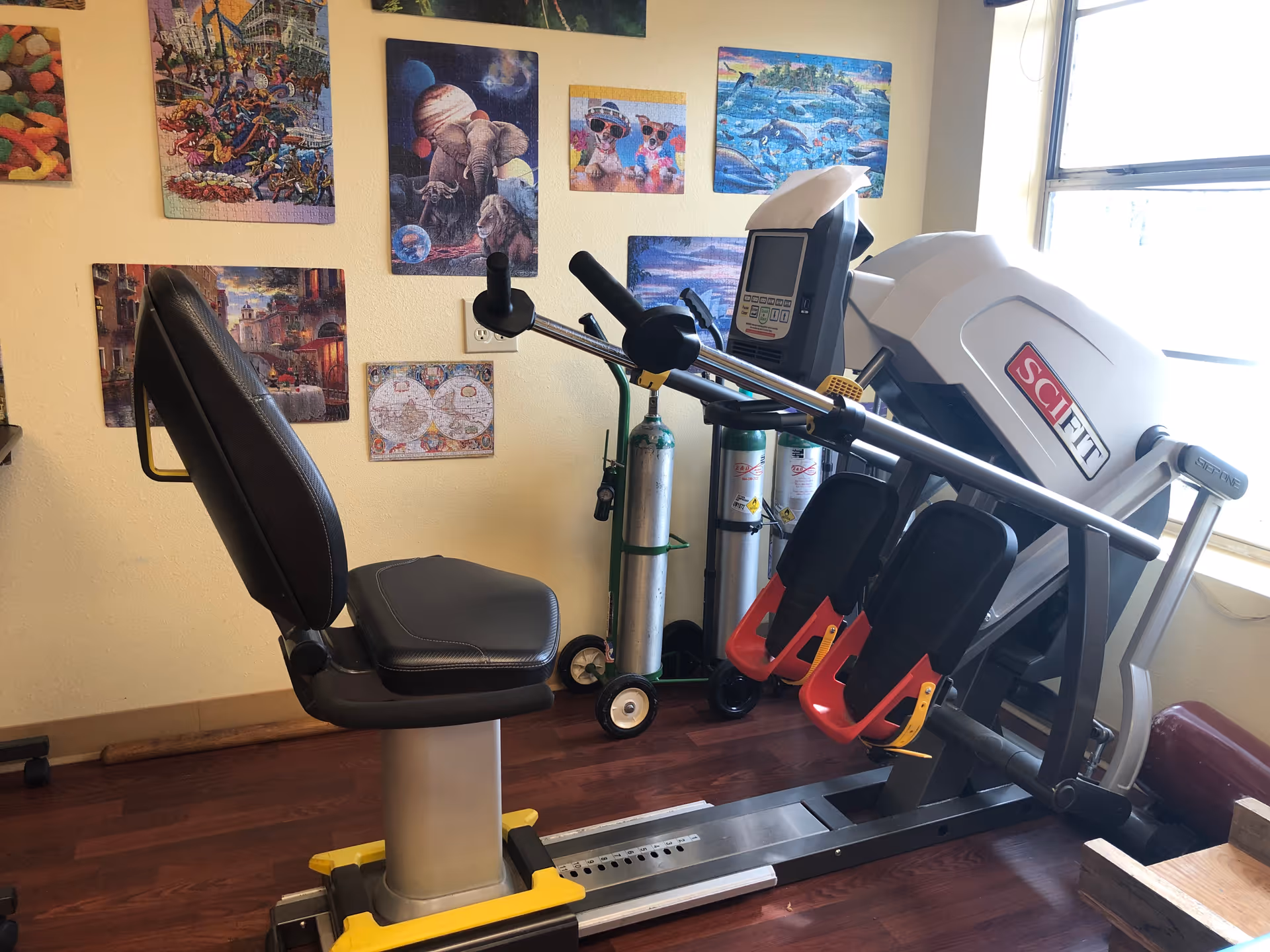 Recumbent exercise machine with oxygen tanks and colorful wall art in a small interior fitness room.