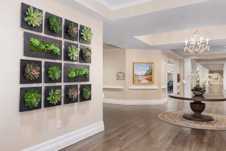 Interior hallway of a senior living facility with a wall display of framed succulent plants on the left, a round wooden table with a flower arrangement on a circular rug in the center, a chandelier hanging from the ceiling, and a framed landscape painting on the far wall.