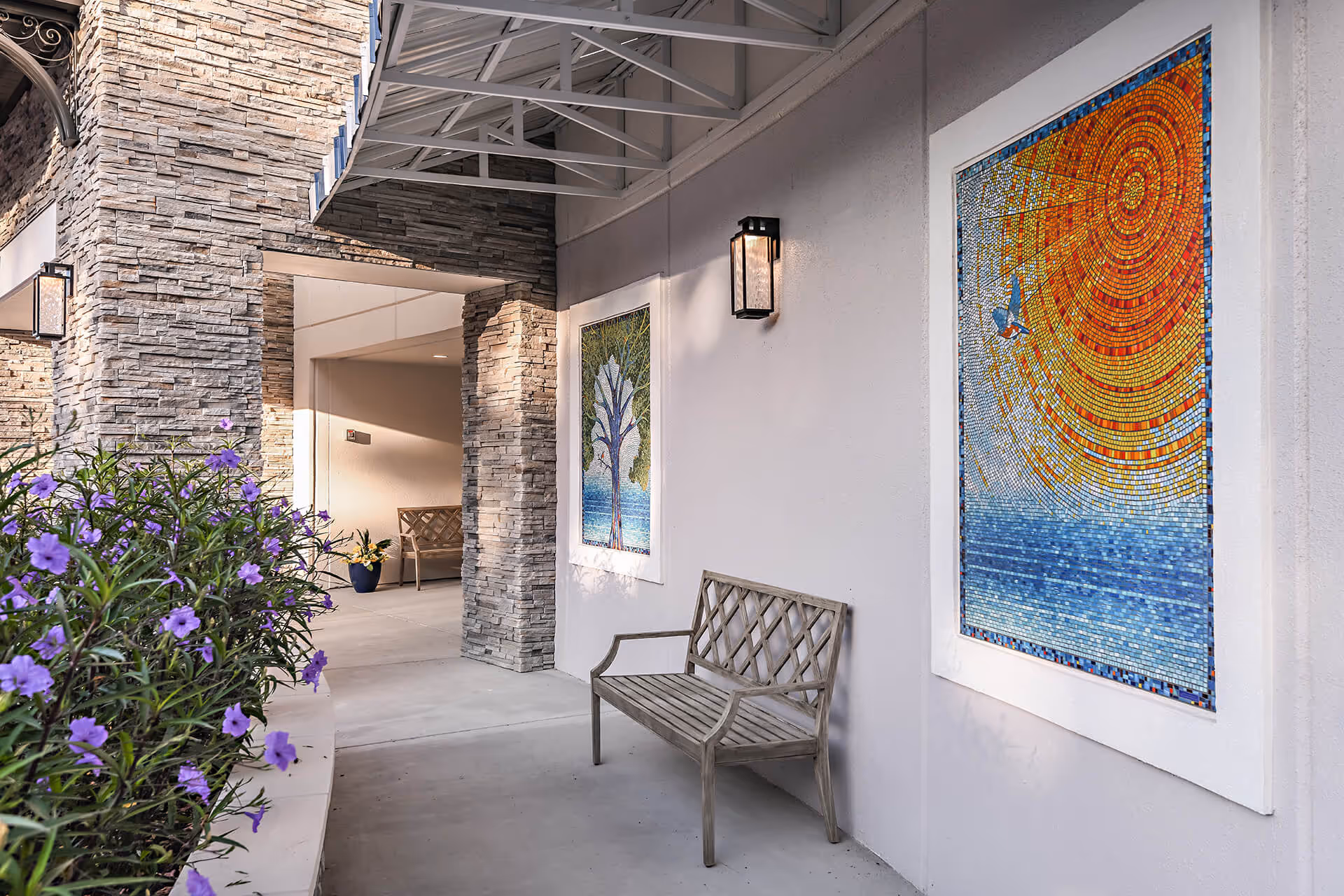 Outdoor covered walkway with stone pillars and a wooden bench. Two colorful mosaic artworks are mounted on the wall, one depicting a tree and the other a sun with a bird flying over water. Purple flowers are planted along the left side of the walkway.