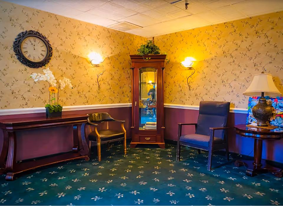 A cozy sitting area with floral wallpaper and green carpet featuring a floral pattern. The room has a wooden console table with a white orchid plant, a black leather chair, a wooden cabinet with glass doors, a blue upholstered armchair, and a round wooden side table with a decorative lamp. Two wall sconces provide warm lighting, and a large round clock hangs on the wall.