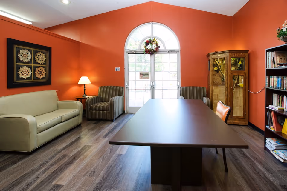 A cozy room with orange walls featuring a beige sofa, two striped armchairs, a wooden table with an orange chair, a floor lamp, a wooden cabinet with glass doors, a bookshelf filled with books, and a large arched window with a wreath hanging on it.