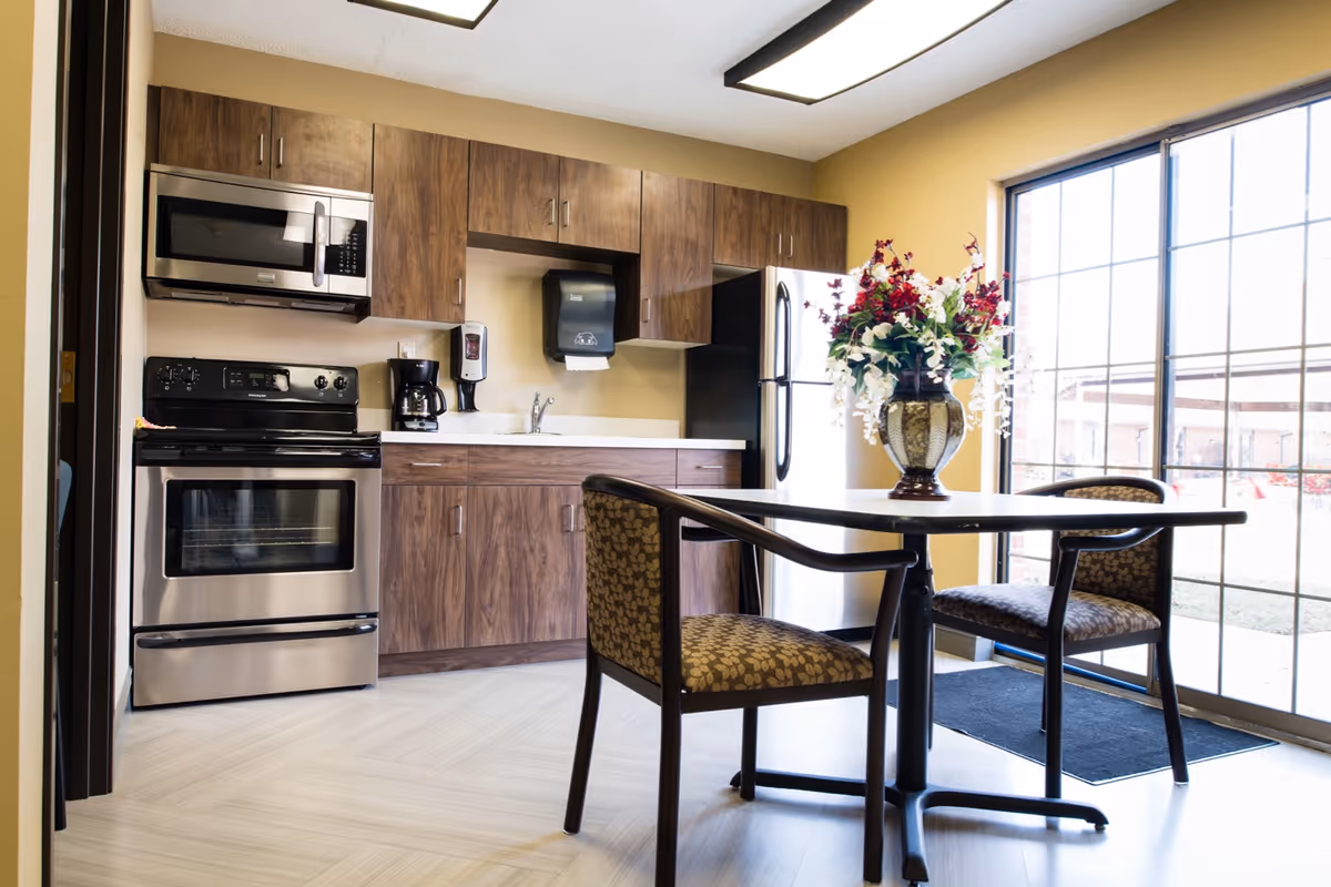 A bright kitchen area with wooden cabinets, a stainless steel oven and microwave, a coffee maker, and a refrigerator. In front of a large window with sliding glass doors, there is a small dining table with two patterned chairs and a decorative vase with flowers on the table.