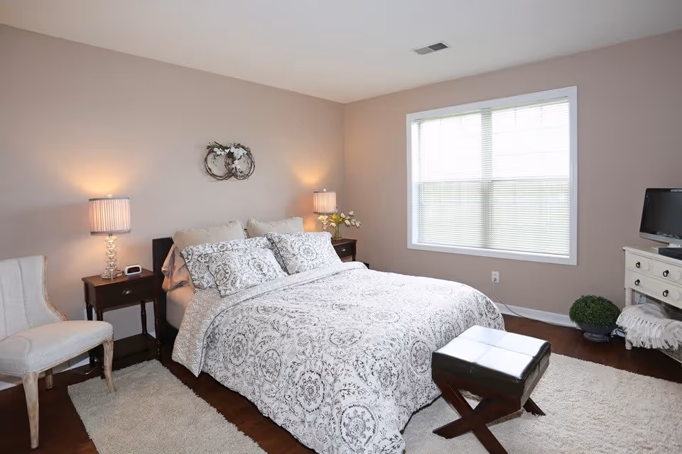A bright bedroom with a large bed covered in a patterned white and gray comforter and matching pillows. There are two bedside tables with lamps on either side of the bed, a beige upholstered chair to the left, a small ottoman at the foot of the bed, a window with blinds on the right wall, and a small TV on a white dresser with a plant and a blanket underneath.