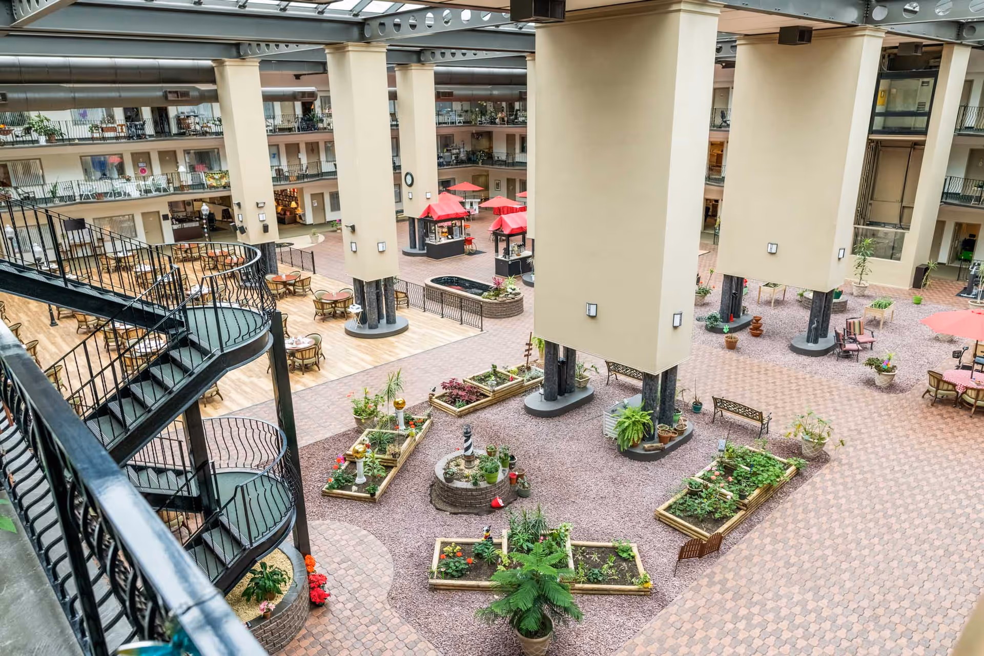 Indoor atrium area of a senior living facility with multiple floors of rooms overlooking a central courtyard. The courtyard features raised garden beds with plants, seating areas with tables and chairs, red umbrellas, and a spiral staircase leading to the upper floors. Large support columns and an elevator are visible, along with decorative plants and benches.
