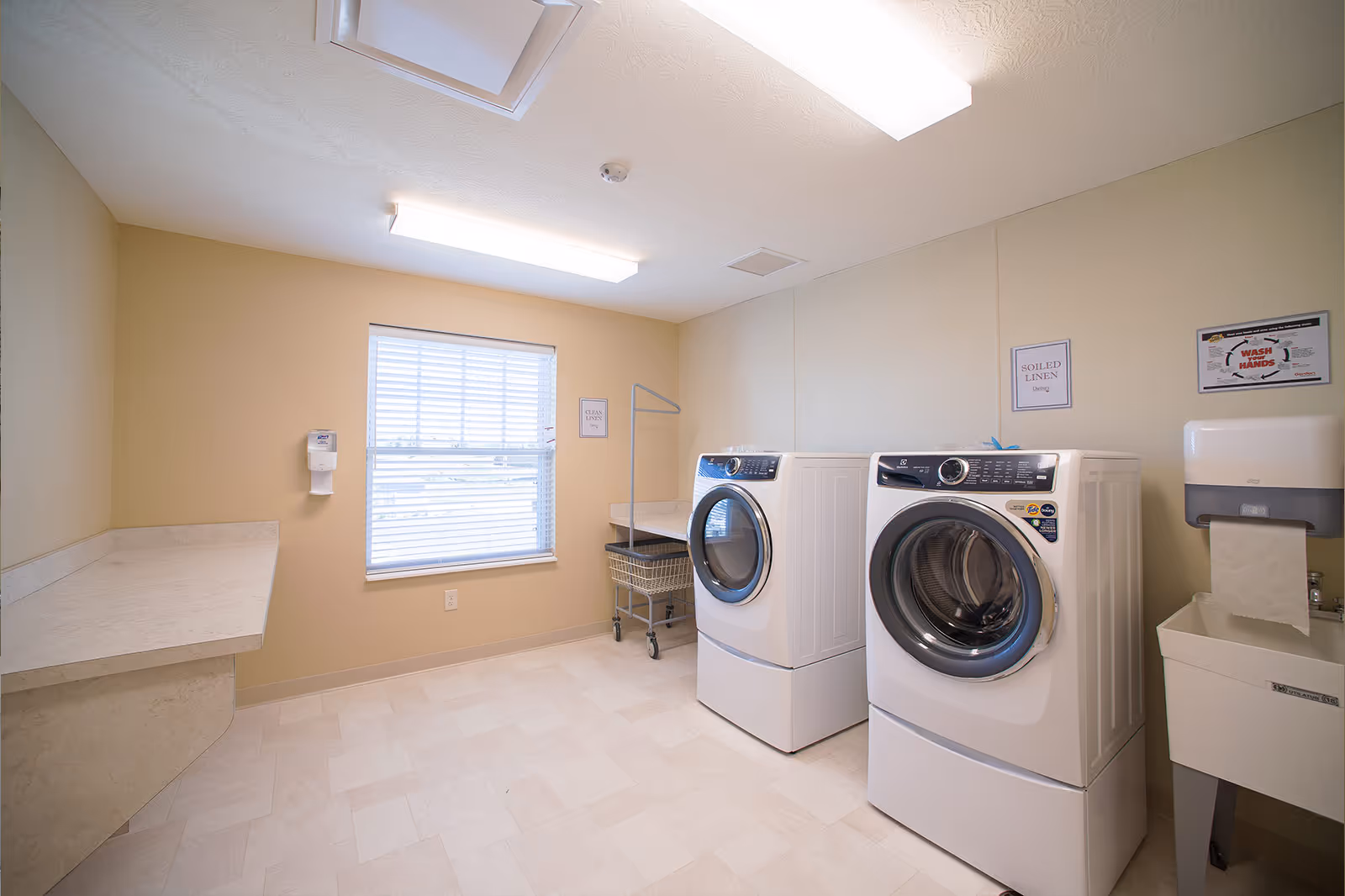 A clean laundry room with two front-loading washing machines, a laundry cart, a countertop for folding clothes, a window with blinds, and signs on the wall indicating areas for clean and soiled linen. There is also a paper towel dispenser and a utility sink.
