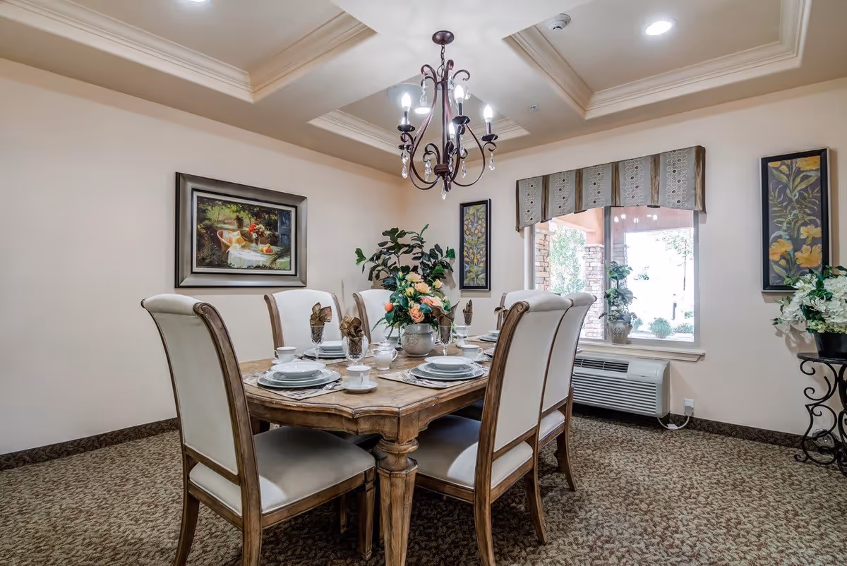 A well-lit dining room with a wooden dining table set for six people. The table is decorated with a floral centerpiece and place settings including plates, cups, and napkins. The room features a chandelier hanging from a tray ceiling, carpeted floor, framed artwork on the walls, a window with a valance, and an air conditioning unit beneath the window.