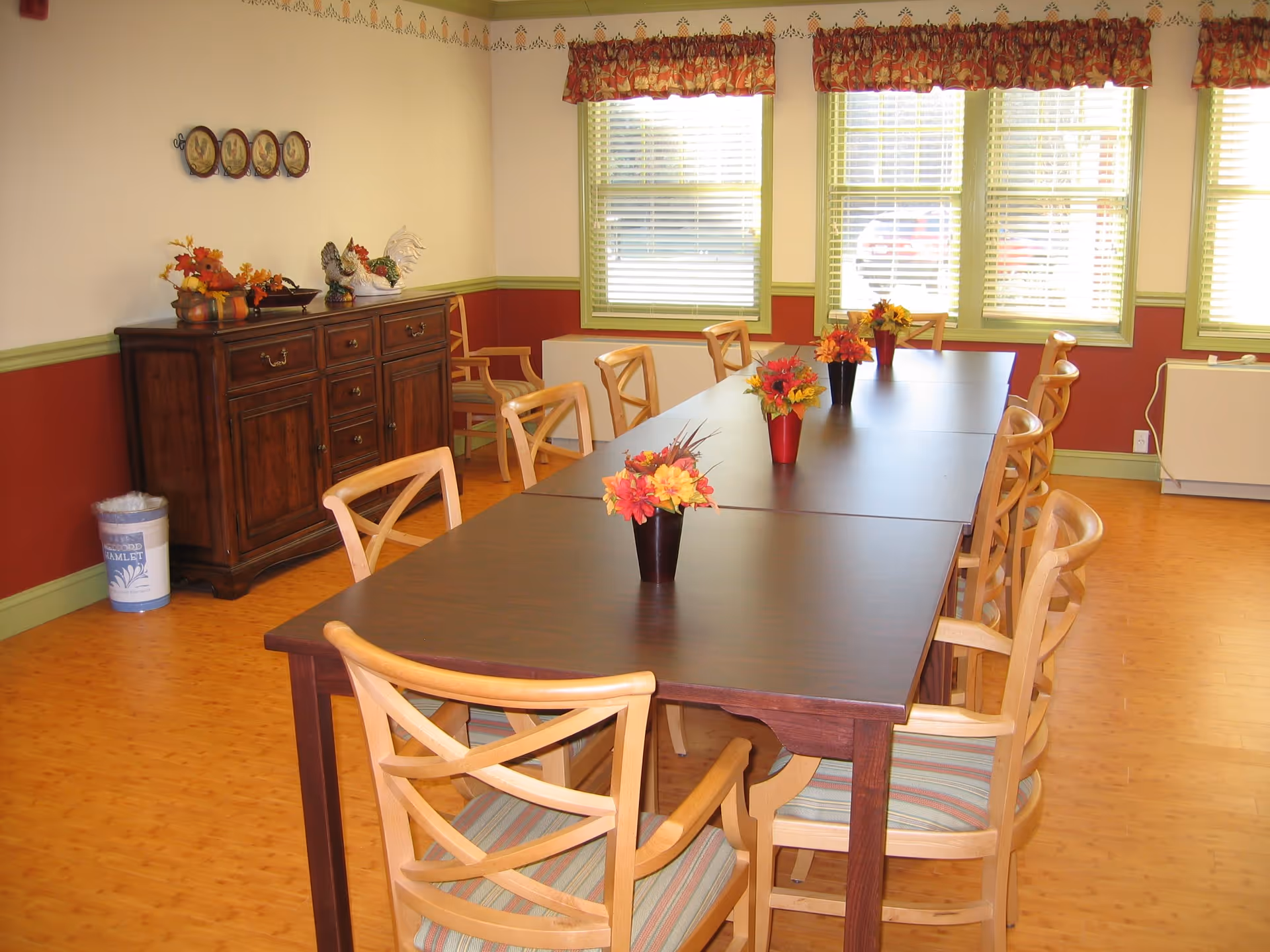 A dining room with a long dark wooden table surrounded by wooden chairs with striped cushions. The table has small vases with colorful artificial flowers. The room has light-colored walls with a red lower half and green trim. There is a wooden sideboard with decorative items and a trash bin in the corner. Three large windows with blinds and floral valances let in natural light.
