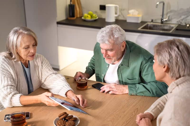 Three elderly people sitting around a wooden table in a kitchen area, looking at a tablet device. There are glasses of tea and a plate of cookies on the table. The background shows a countertop with a sink, a kettle, a tissue box, and a bowl of green apples.