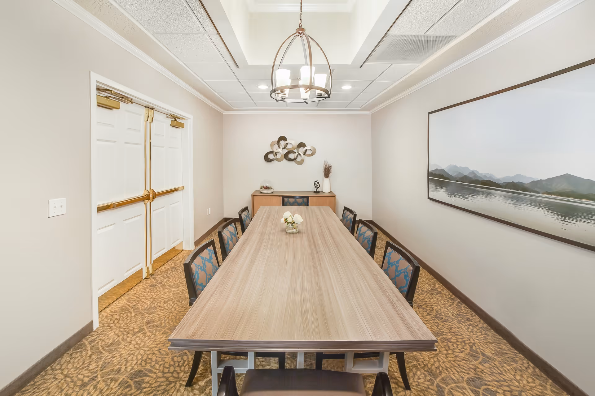 A long rectangular wooden table with eight chairs in a well-lit room. The room has beige walls, a patterned carpet, a modern chandelier hanging from the ceiling, a large landscape painting on the right wall, and a sideboard with decorative items against the far wall. Double white doors with gold handles are on the left side.