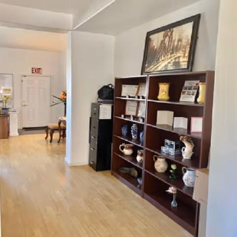Interior hallway area with a wooden shelving unit displaying vases and awards, light wood flooring, and an exit door in the background.