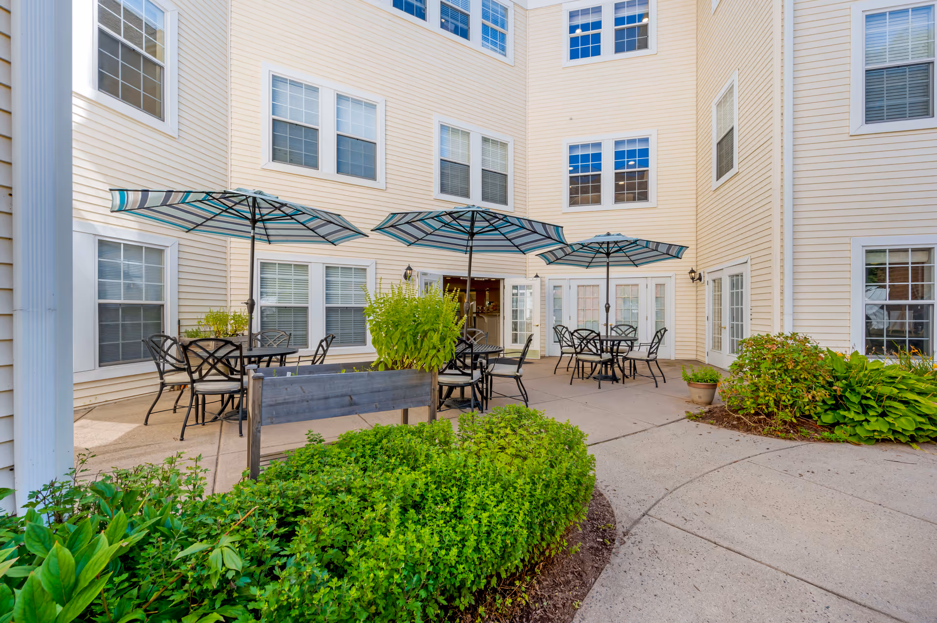 Outdoor courtyard area of a senior living facility with several black metal tables and chairs under blue and white striped umbrellas. The courtyard is surrounded by a beige multi-story building with many windows. There are green shrubs and plants along the edges of the paved area.