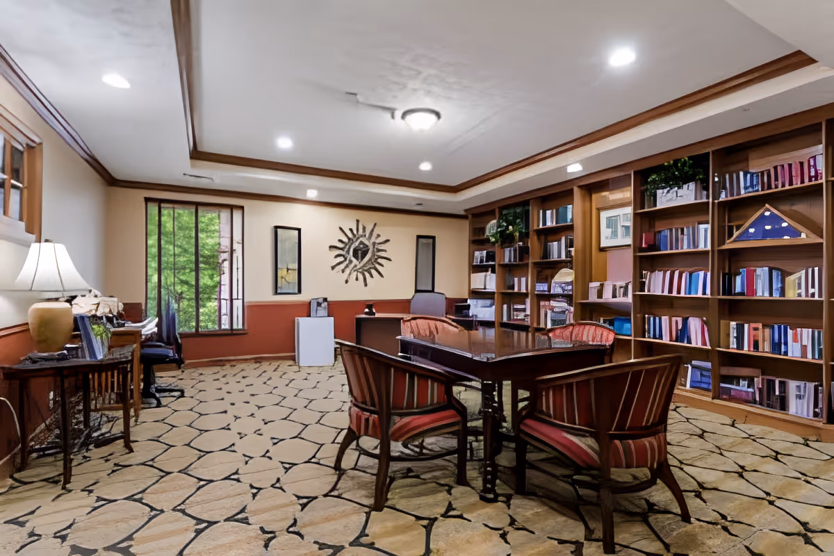 A cozy library or study room with a wooden table surrounded by four striped upholstered chairs. The room features built-in wooden bookshelves filled with books and decorative items. There is a window with a view of greenery, a small table with a lamp and flowers, and a decorative sun-shaped wall art. The ceiling has recessed lighting and a central light fixture.