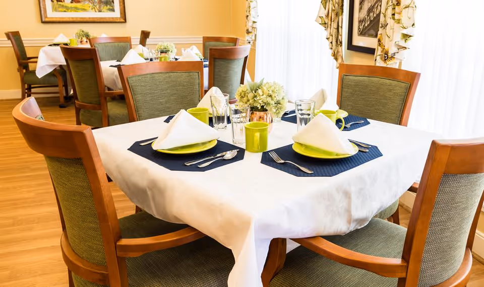 A dining area with wooden chairs featuring green cushions around tables covered with white tablecloths. Each table is set with green plates, folded white napkins, silverware, glasses, and green mugs. There are floral centerpieces on the tables and a framed picture on the wall in the background.