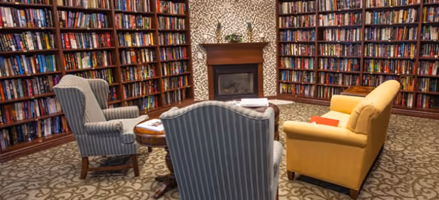 A cozy library room with tall wooden bookshelves filled with books lining the walls. In the center, there is a small round wooden table surrounded by two striped armchairs and a yellow sofa. A fireplace with a wooden mantle is set against the far wall, with decorative items on top. The floor is carpeted with a patterned design.