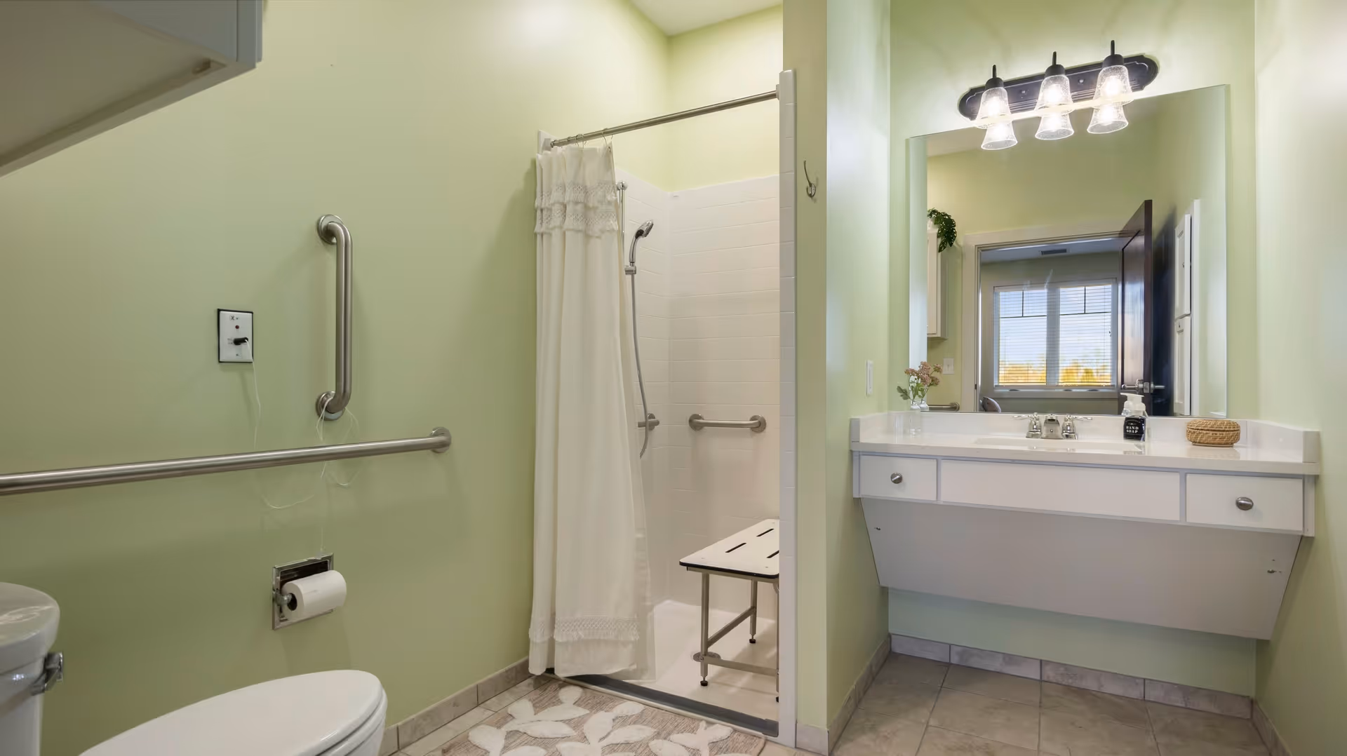 Accessible bathroom with a roll-in shower and shower chair, grab bars, toilet, and a vanity with mirror and lights.