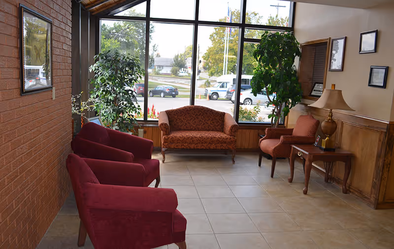 A cozy seating area inside a facility with large windows showing a parking lot outside. The room has three red armchairs, one patterned loveseat, a patterned armchair, a wooden side table with a lamp, and two large green plants near the windows. The walls are a combination of brick and wood paneling.